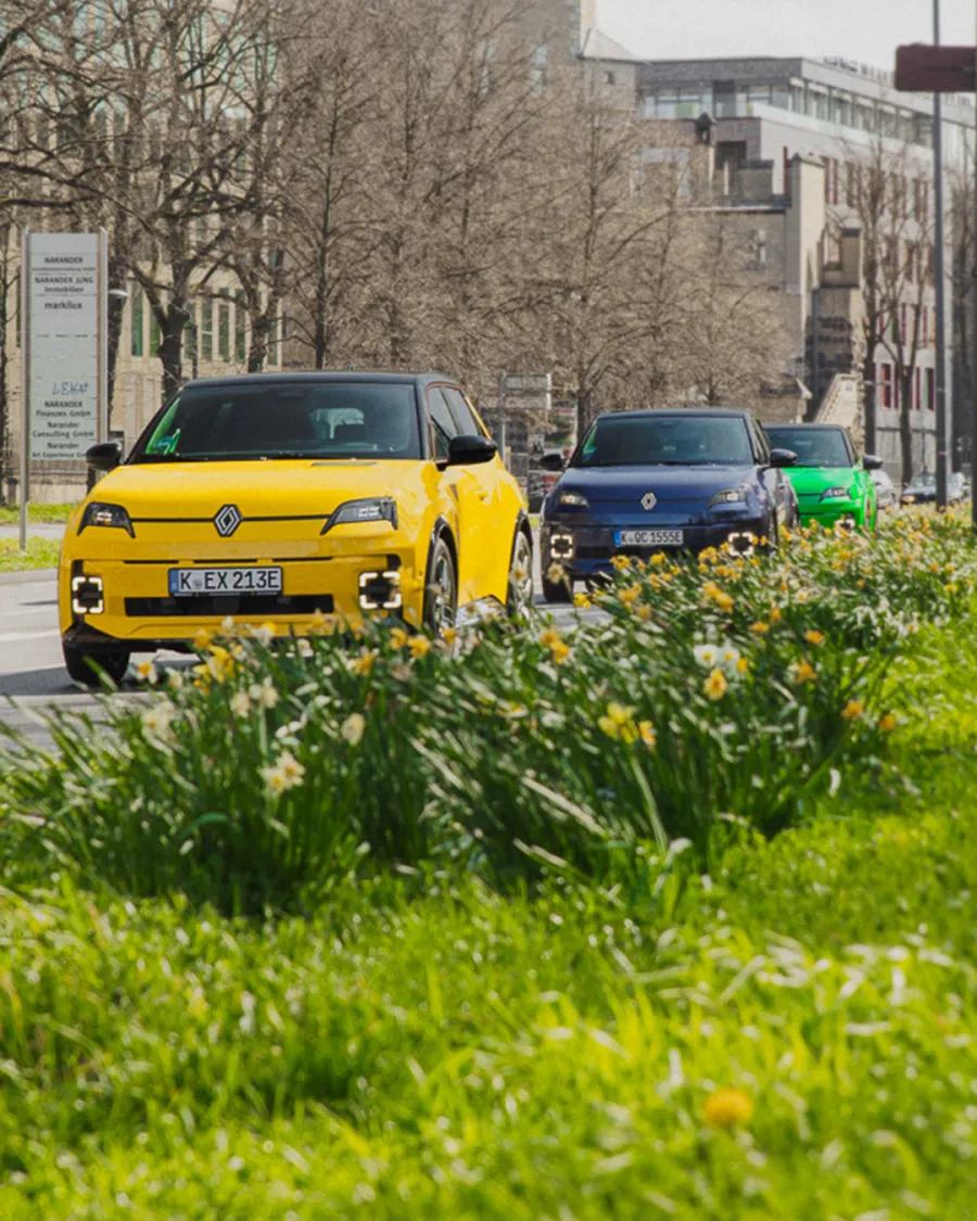 Gelber und grüner Renault 5 E-Tech in Fahrt auf einer Kölner Straße mit Parkbepflanzung im Hintergrund